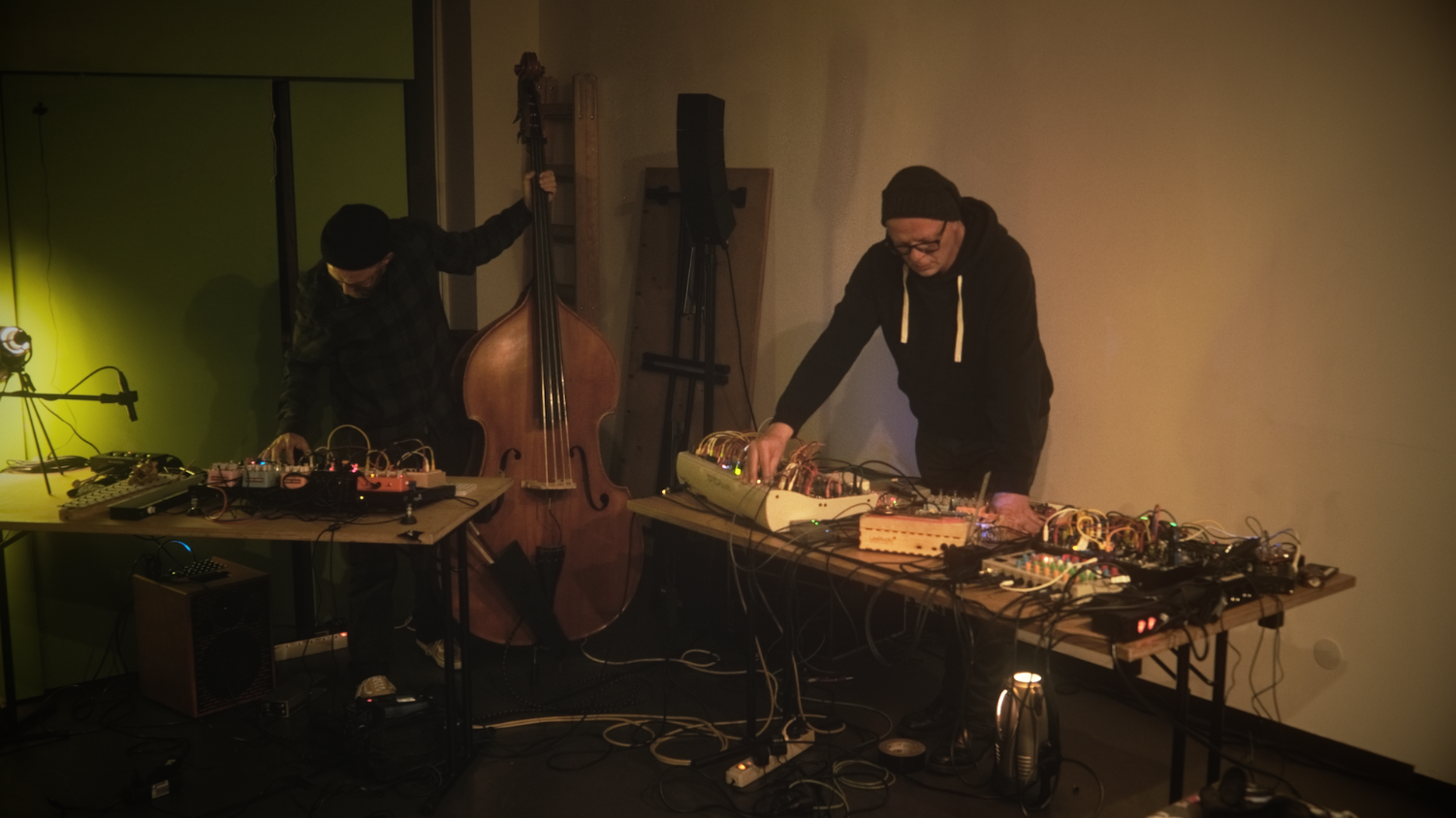 Zwei Klangkünstler bei einer Live-Performance mit Kontrabass, Modular-Synthesizern und Effektgeräten. Die Szene ist in warmes Licht getaucht, eine intime Atmosphäre zwischen analogem Sound und experimenteller Elektronik.